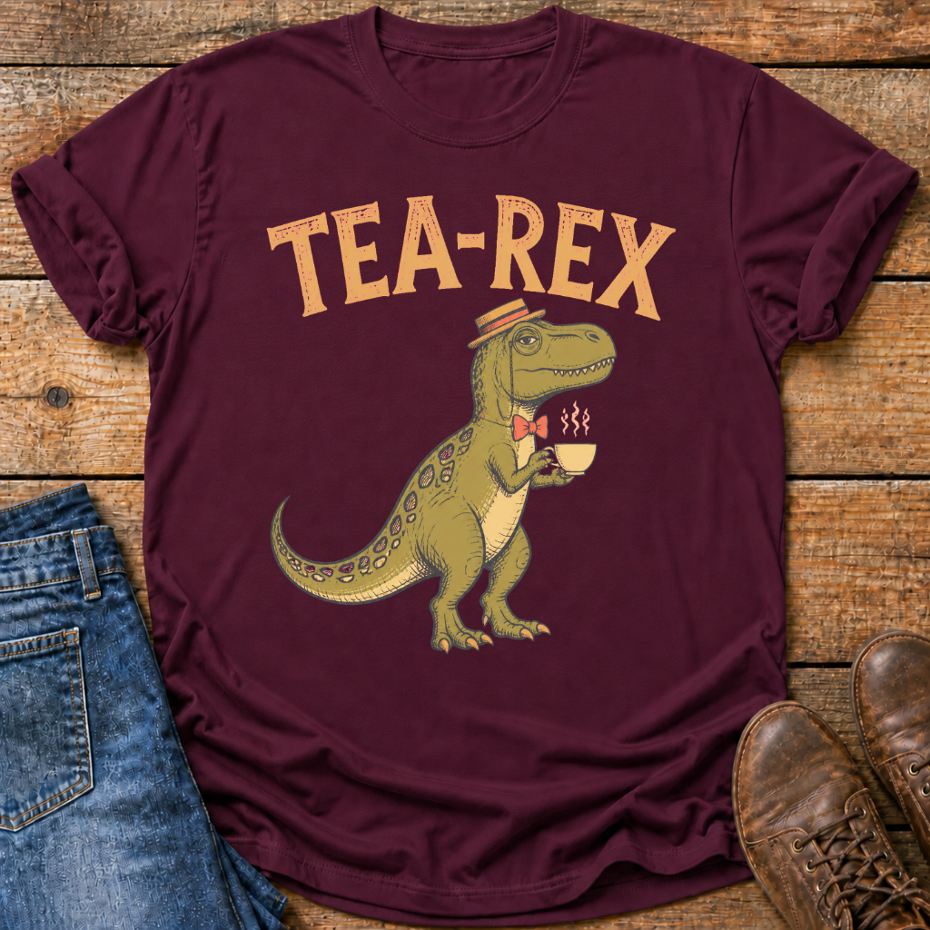 A maroon short-sleeve graphic T-shirt laid flat on a rustic wood surface, styled with blue jeans on the left and brown shoes on the right. The shirt features a large tan 'TEA-REX' text and a cartoon green dinosaur wearing a hat and bow tie while holding a teacup.