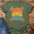 An olive green graphic t-shirt with the text 'BAKERS GONNA BAKE' in retro orange, yellow, and blue lettering, styled in a flat lay on a wooden surface next to blue jeans and brown shoes.