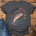 A dark charcoal gray t-shirt with a colorful graphic of a jumping rainbow trout and the text 'CATCH ME OUTSIDE' in blue and red, styled in a flat lay on a rustic wooden background with blue jeans and brown boots.