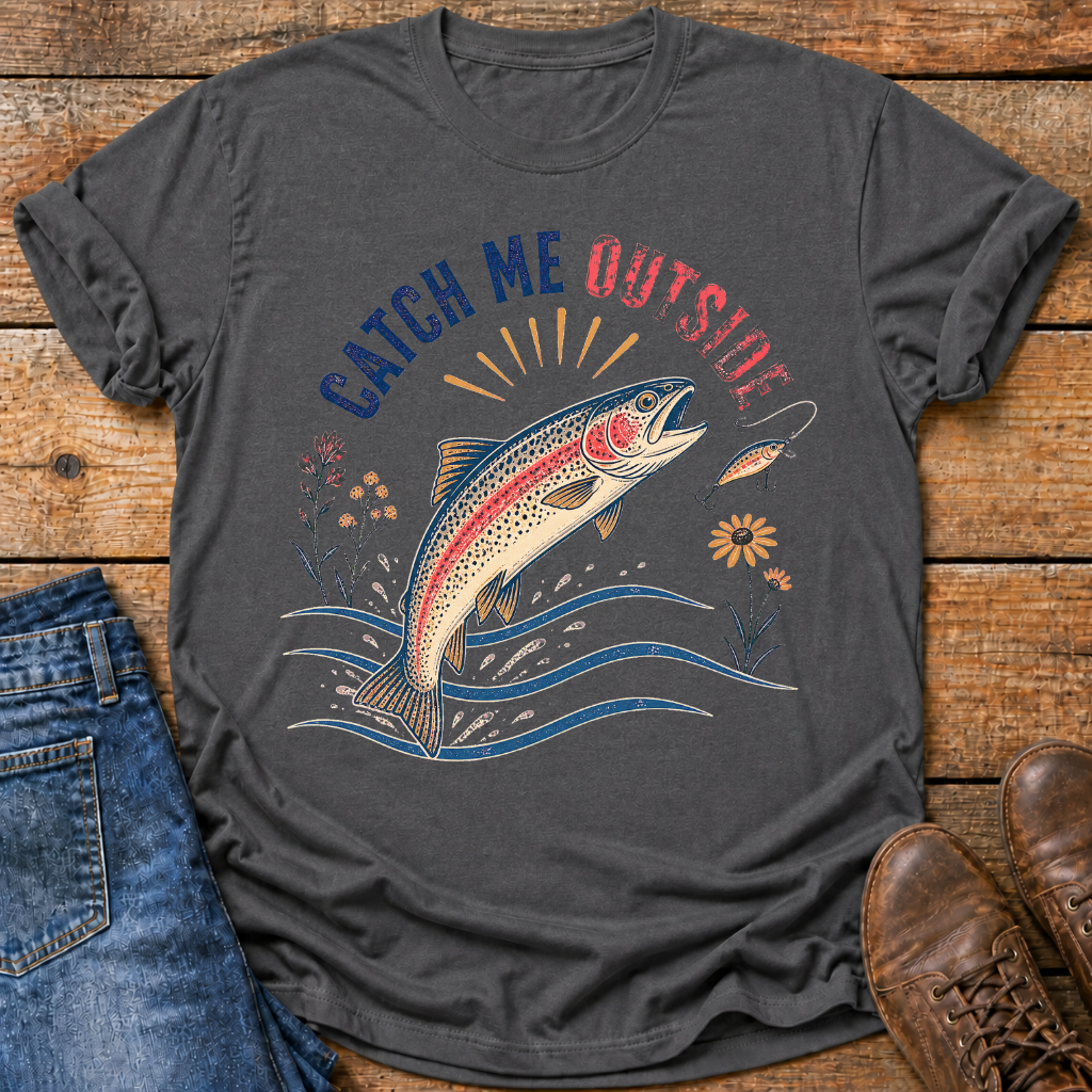 A dark charcoal gray t-shirt with a colorful graphic of a jumping rainbow trout and the text 'CATCH ME OUTSIDE' in blue and red, styled in a flat lay on a rustic wooden background with blue jeans and brown boots.