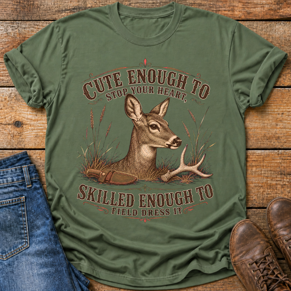 An olive green graphic t-shirt laid flat on a rustic wooden surface, styled with a pair of blue jeans and brown leather boots. The t-shirt features a detailed graphic of a deer with antlers and the text 'CUTE ENOUGH TO STOP YOUR HEART.