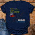 A navy blue graphic t-shirt laid flat on a rustic wooden surface, styled with folded blue jeans on the left and brown leather boots on the right. The t-shirt features a distressed text graphic reading 'NOT ALL WHO WANDER ARE LOST SOME ARE TRACKING' in olive green, white, and red lettering, accompanied by a trail of footprints.