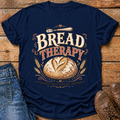A navy blue graphic t-shirt laid flat on a rustic wooden surface, styled with blue jeans on the left and brown leather shoes on the right. The t-shirt features a retro-style graphic with the words 'BREAD THERAPY' in cream and orange-brown lettering, an illustration of a crusty loaf of bread, wheat stalks, and a small knife.