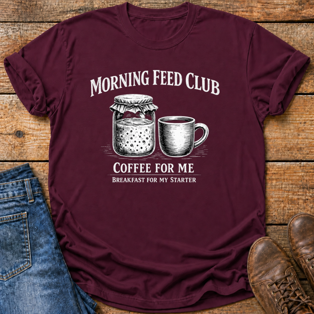 A maroon short-sleeve graphic T-shirt laid flat on a rustic wood surface, styled with blue jeans on the left and brown shoes on the right. The shirt has a white printed design reading “MORNING FEED CLUB” above an illustration of a jar and mug, and “COFFEE FOR ME / BREAKFAST FOR MY STARTER” below.