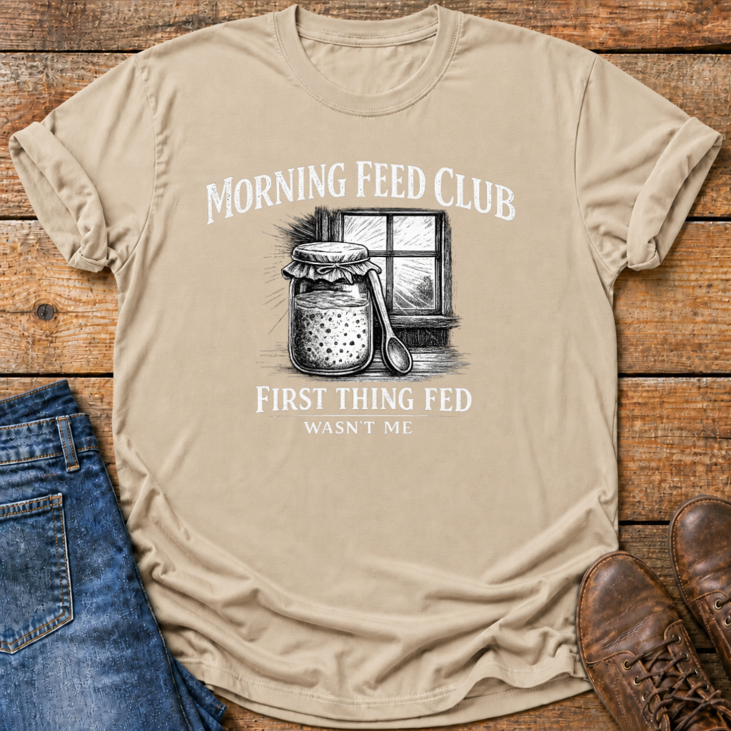 A beige graphic t-shirt laid flat on a rustic wooden background, styled with blue jeans and brown leather shoes. The t-shirt features a black and white illustration of a jar of pickles next to a window, with the text 'MORNING FEED CLUB' above and 'FIRST THING FED WASN'T ME' below.
