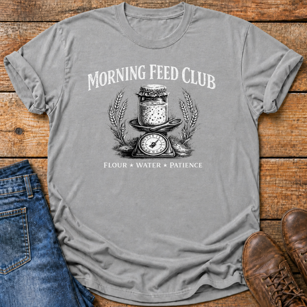 A heather gray short-sleeve t-shirt with a vintage-style black graphic reading 'MORNING FEED CLUB' and 'FLOUR * WATER * PATIENCE' around an illustration of a flour jar, scale, and wheat ears. The shirt is styled in a flat lay on a rustic wooden background with blue jeans and brown leather shoes partially visible.