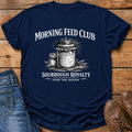 A navy blue graphic t-shirt laid flat on a rustic wooden surface, styled with blue jeans on the left and brown leather shoes on the right. The t-shirt features a white graphic print of a crowned jar of sourdough bread with the text 'MORNING FEED CLUB' and 'SOURDOUGH ROYALTY' and 'FEED THE QUEEN'.