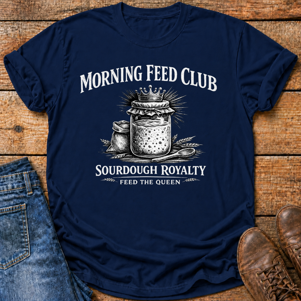 A navy blue graphic t-shirt laid flat on a rustic wooden surface, styled with blue jeans on the left and brown leather shoes on the right. The t-shirt features a white graphic print of a crowned jar of sourdough bread with the text 'MORNING FEED CLUB' and 'SOURDOUGH ROYALTY' and 'FEED THE QUEEN'.