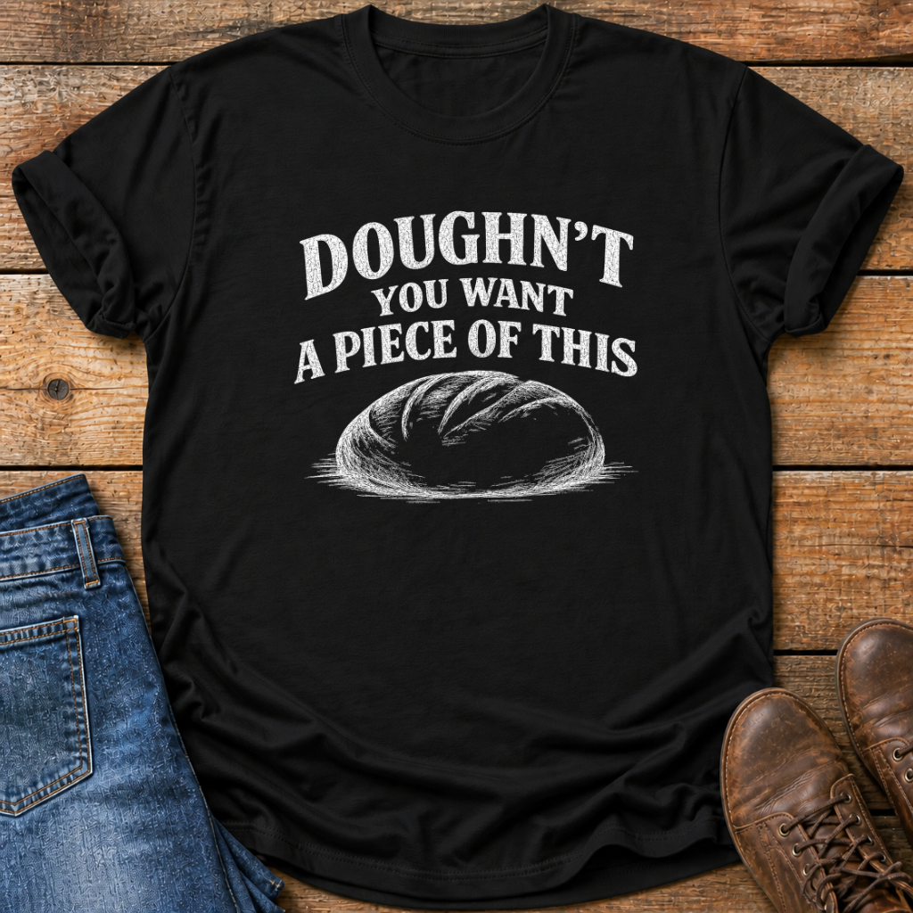A black short-sleeve t-shirt with a white graphic print that reads 'DOUGHN'T YOU WANT A PIECE OF THIS' above a sketch of a loaf of bread. The shirt is styled in a flat-lay on a rustic wooden background, partially covered by blue jeans on the left and brown leather shoes on the right.