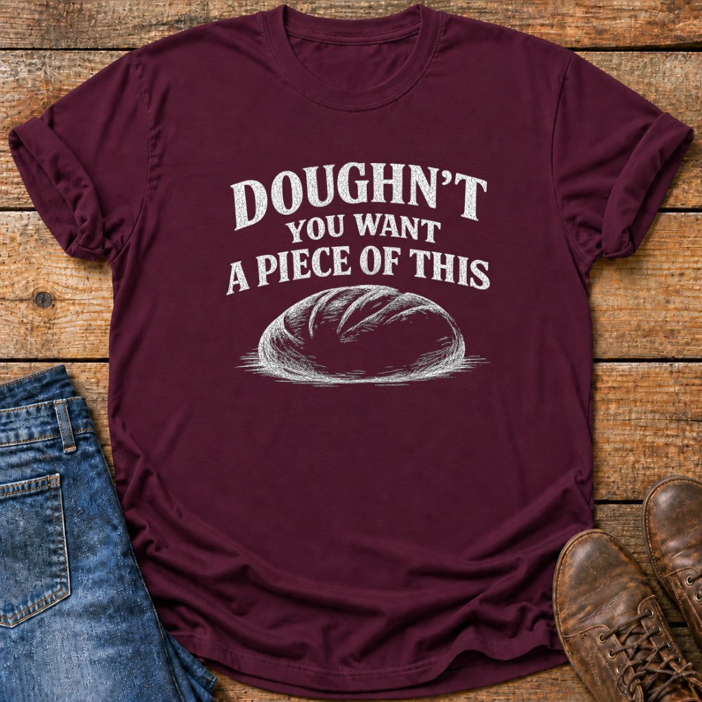 A maroon short-sleeve t-shirt with a white graphic print that reads 'DOUGHN'T YOU WANT A PIECE OF THIS' above a sketch of a loaf of bread. The shirt is styled in a flat-lay on a rustic wooden background, partially covered by blue jeans on the left and brown leather shoes on the right.