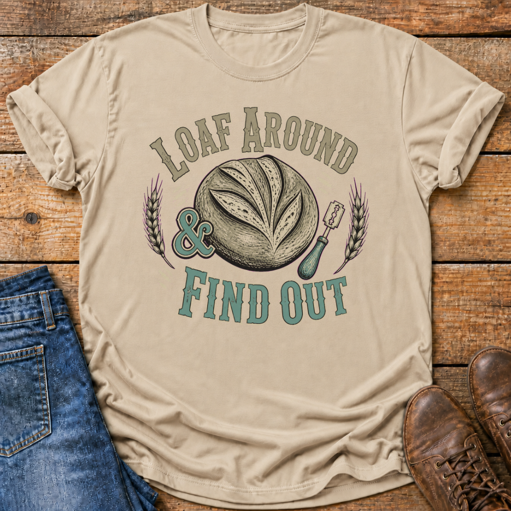 A beige graphic t-shirt laid flat on a rustic wooden surface, styled with blue jeans on the left and brown leather shoes on the right. The t-shirt features a retro-style graphic of a sliced loaf of bread with the text 'LOAF AROUND & FIND OUT' in teal and brown.