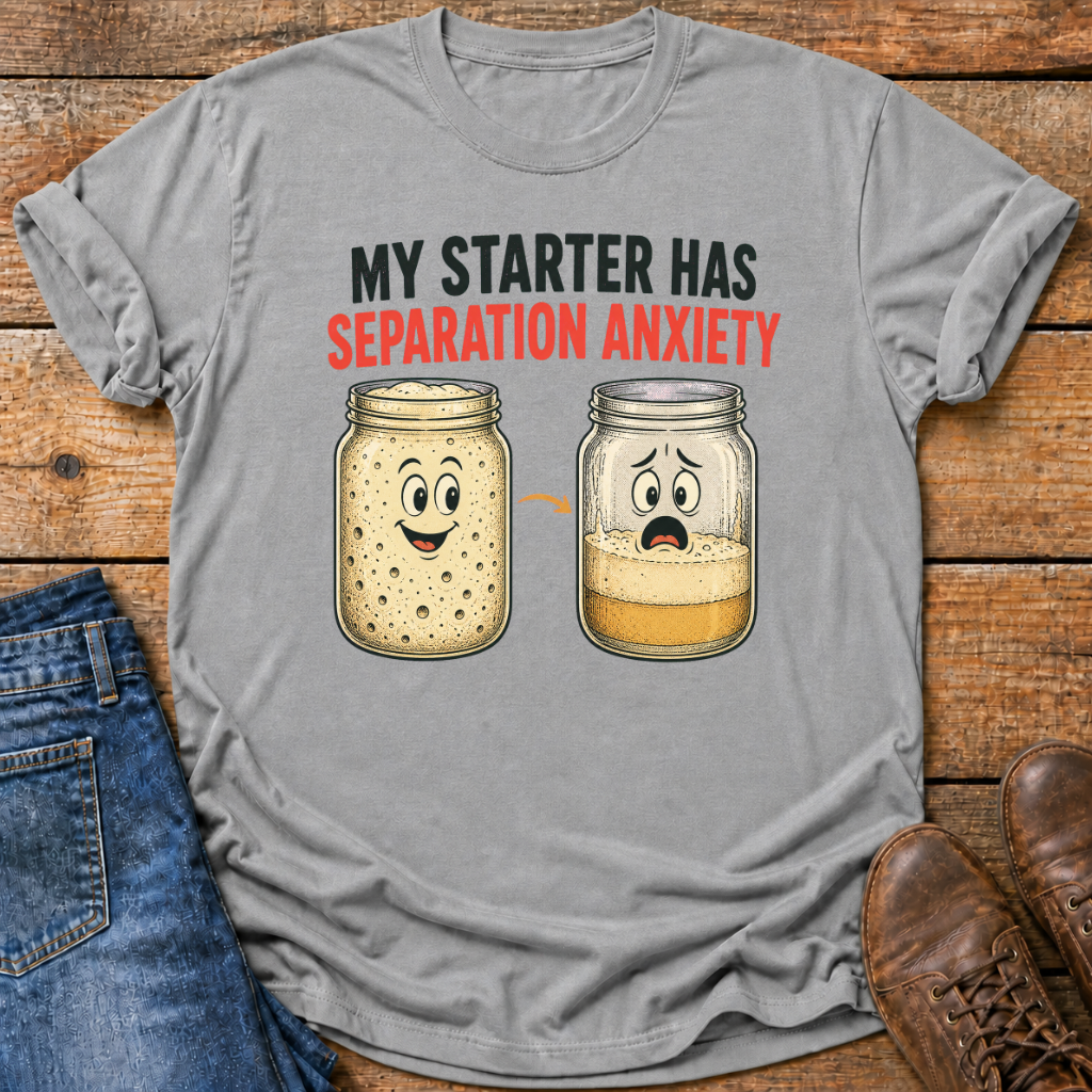A gray graphic t-shirt laid flat on a rustic wooden background, styled with blue jeans and brown leather shoes. The t-shirt features a humorous graphic with the text 'MY STARTER HAS SEPARATION ANXIETY' above two cartoon mason jars showing yeast separation.