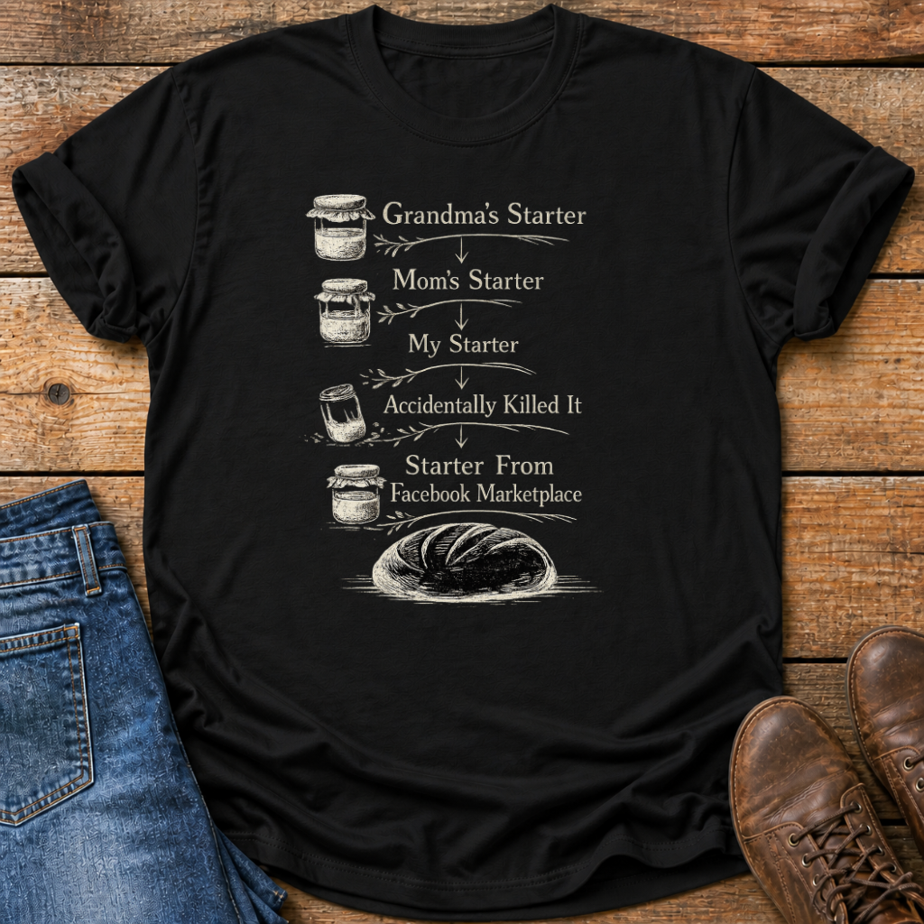 A black short-sleeve graphic T-shirt laid flat on a rustic wooden surface, styled with blue jeans on the left and brown leather shoes on the right. The shirt features a white printed humorous sequence starting with 'Grandma's Starter' and ending with 'Starter From Facebook Marketplace' accompanied by illustrations of jars and a loaf of bread.