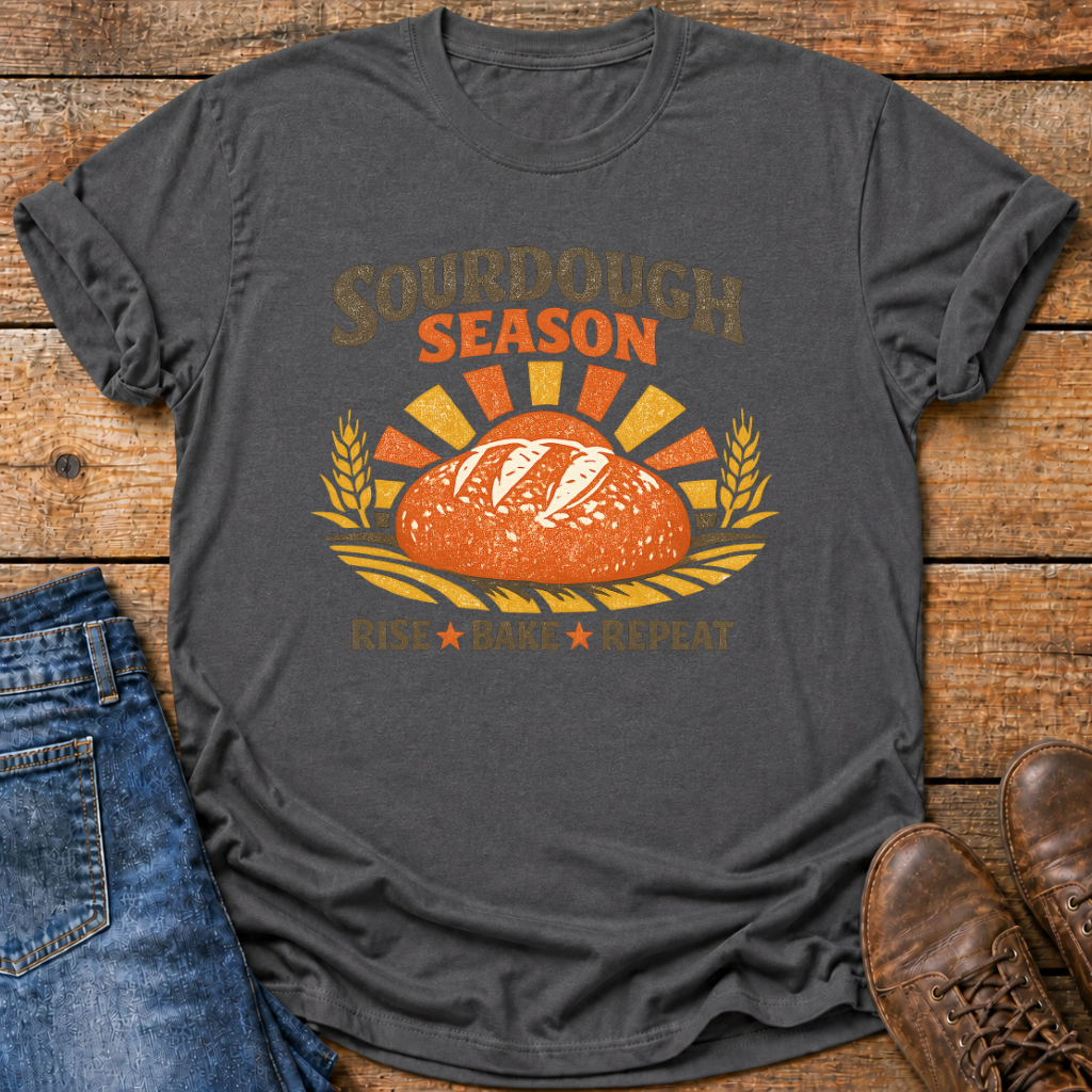A dark charcoal gray graphic t-shirt laid flat on a rustic wooden surface, styled with blue jeans and brown leather shoes. The t-shirt features a retro-style graphic of a sourdough bread loaf with wheat ears and the text 'SOURDOUGH SEASON' and 'Rise Bake Repeat'.