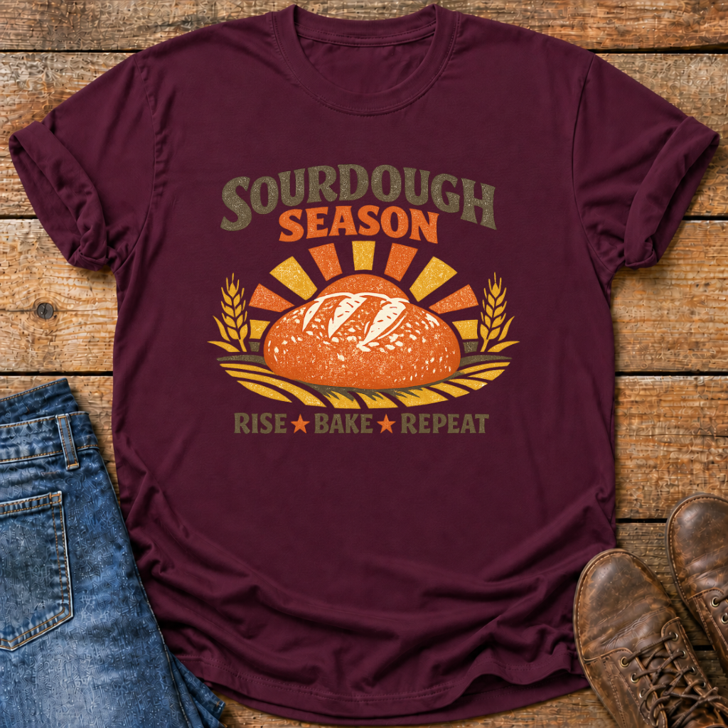 A maroon graphic t-shirt laid flat on a rustic wooden surface, styled with blue jeans and brown leather shoes. The t-shirt features a retro-style graphic of a sourdough bread loaf with wheat ears and the text 'SOURDOUGH SEASON' and 'Rise Bake Repeat'.