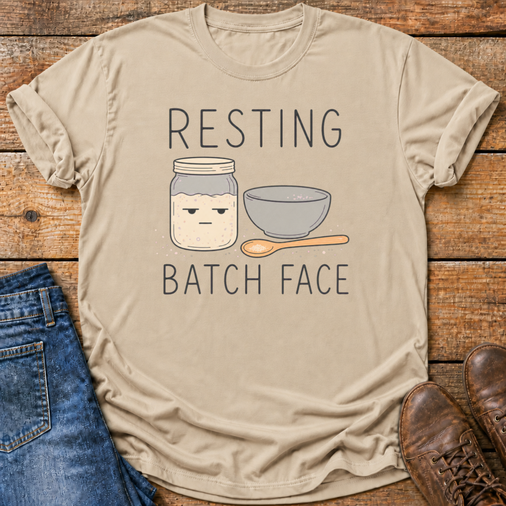 A beige graphic t-shirt laid flat on a rustic wooden surface, styled with blue jeans on the left and brown shoes on the right. The t-shirt features a cartoon illustration of a grumpy jar with a face, a gray bowl, and a wooden spoon, with the text 'RESTING BATCH FACE' printed above and below the graphic.