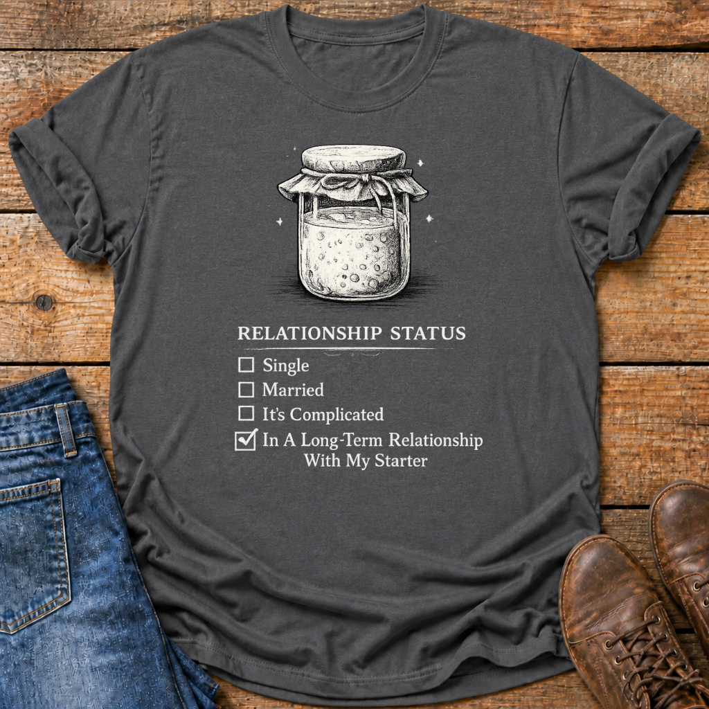 A dark charcoal gray graphic t-shirt laid flat on a rustic wooden surface, styled with blue jeans on the left and brown leather shoes on the right. The t-shirt features a black line art illustration of a glass jar with a bow on the lid, above a text graphic that reads 'RELATIONSHIP STATUS' with checkboxes for 'Single', 'Married', 'It's Complicated', and 'In A Long-Term Relationship With My Starter' checked.