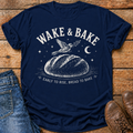 A navy blue graphic t-shirt laid flat on a rustic wooden surface, styled with a pair of blue jeans on the left and brown leather shoes on the right. The t-shirt features a white graphic of a bird perched on a loaf of bread under a crescent moon and stars, with the text 'WAKE & BAKE' and 'EARLY TO RISE, BREAD TO BAKE'.