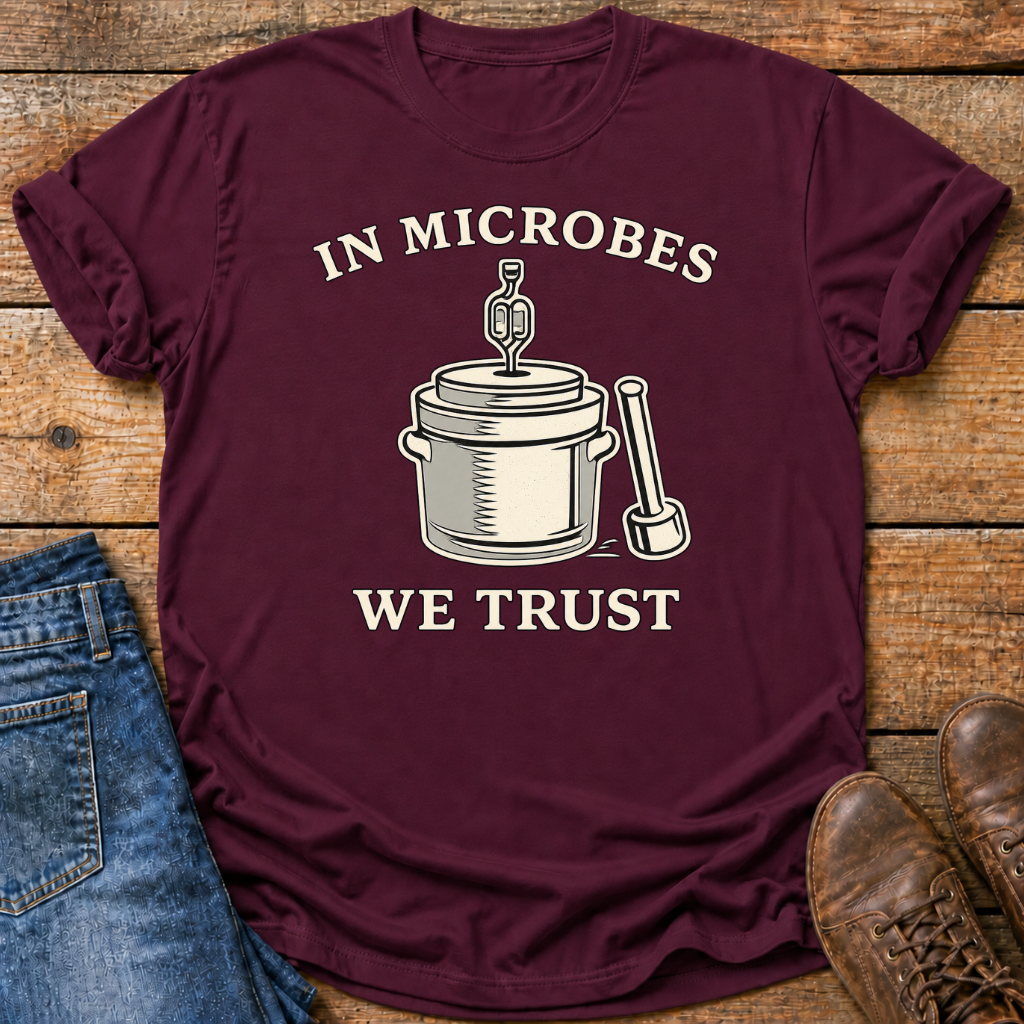A maroon short-sleeve graphic T-shirt laid flat on a rustic wooden surface, styled with blue jeans on the left and brown shoes on the right. The shirt has a cream-colored graphic of a laboratory pot and pipette with the text 'IN MICROBES WE TRUST'.