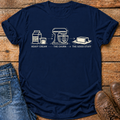A navy blue graphic t-shirt laid flat on a rustic wooden surface, styled with blue jeans, brown leather shoes, and a black coffee mug. The t-shirt features a white line-art graphic of heavy cream, a stand mixer, and butter with the text 'HEAVY CREAM .