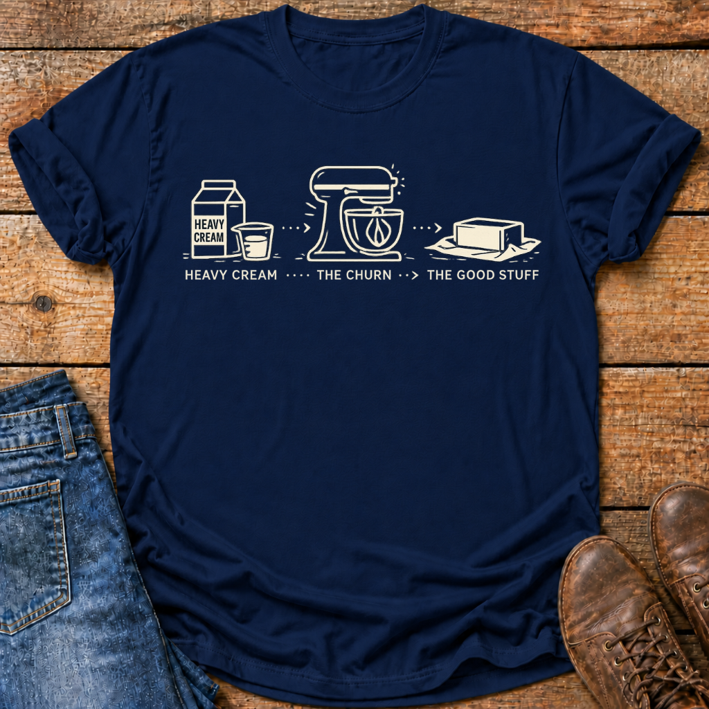 A navy blue graphic t-shirt laid flat on a rustic wooden surface, styled with blue jeans, brown leather shoes, and a black coffee mug. The t-shirt features a white line-art graphic of heavy cream, a stand mixer, and butter with the text 'HEAVY CREAM .