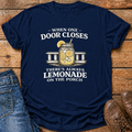 A navy blue graphic t-shirt laid flat on a rustic wooden surface, styled with blue jeans on the left and brown leather shoes on the right. The t-shirt features a graphic of a lemonade mason jar with lemons and the text 'WHEN ONE DOOR CLOSES THERE'S ALWAYS LEMONADE ON THE PORCH'.