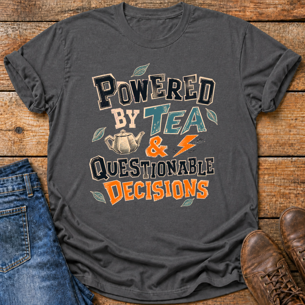 A dark charcoal gray graphic t-shirt laid flat on a rustic wooden background, styled with blue jeans and brown leather boots. The t-shirt features a retro-style graphic that reads 'POWERED BY TEA & QUESTIONABLE DECISIONS' in black, teal, and orange lettering, accompanied by illustrations of a teapot and leaves.