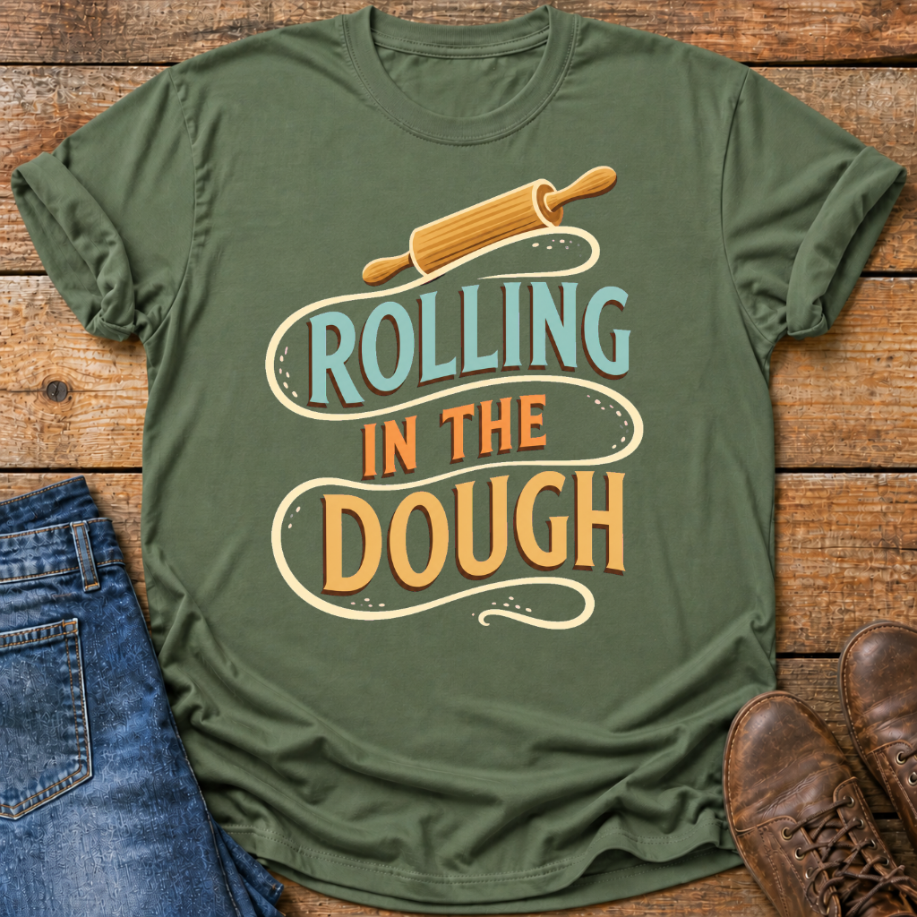 An olive green graphic t-shirt laid flat on a rustic wooden surface, styled with blue jeans on the left and brown leather shoes on the right. The t-shirt features a retro-style graphic with a wooden rolling pin and the text 'ROLLING IN THE DOUGH' in light blue and orange lettering.