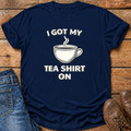 A navy blue graphic t-shirt with the white text 'I GOT MY TEA SHIRT ON' and a white teacup illustration, styled in a flat lay on a rustic wooden background with jeans and brown shoes.