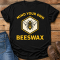 A black short-sleeve graphic T-shirt laid flat on a rustic wood surface, styled with blue jeans on the left and brown shoes on the right. The shirt has a large front graphic with the words “MIND YOUR OWN BEESWAX” in yellow and a central hexagon containing a bee illustration on a cream background.