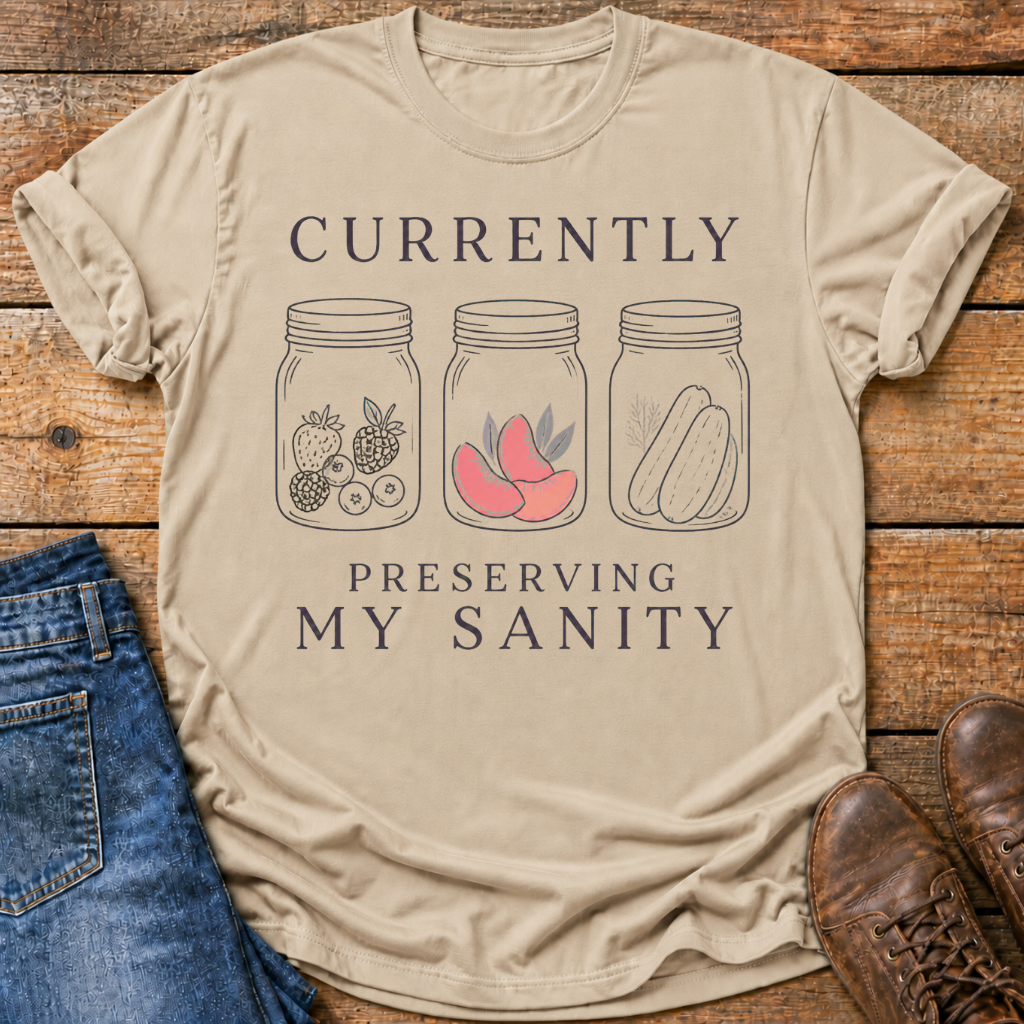 A beige graphic t-shirt laid flat on a rustic wooden surface, styled with blue jeans and brown leather shoes. The t-shirt features a printed design with the text 'CURRENTLY PRESERVING MY SANITY' and illustrations of mason jars containing fruit, peaches, and pickles.