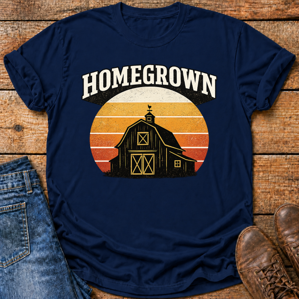 A navy blue graphic t-shirt laid flat on a rustic wooden surface, styled with folded blue jeans on the left and brown leather shoes on the right. The t-shirt features a retro sunset graphic with a black barn silhouette and the word 'HOMEGROWN' in white arched text above it.