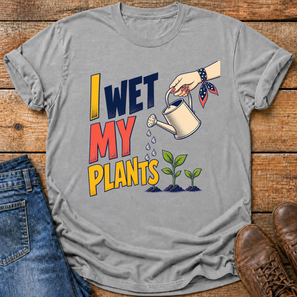 A heather gray graphic t-shirt laid flat on a rustic wooden surface, styled with blue jeans on the left and brown leather shoes on the right. The t-shirt features a colorful graphic that reads 'I WET MY PLANTS' with an illustration of a hand holding a watering can and two small green seedlings.