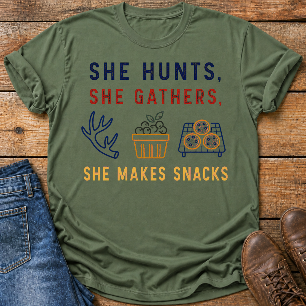 An olive green graphic t-shirt laid flat on a rustic wooden background, styled with blue jeans on the left and brown leather shoes on the right. The t-shirt features the text 'SHE HUNTS, SHE GATHERS, SHE MAKES SNACKS' in navy blue and red lettering, accompanied by simple illustrations of a deer antler, a basket of apples, and a tray of cookies.