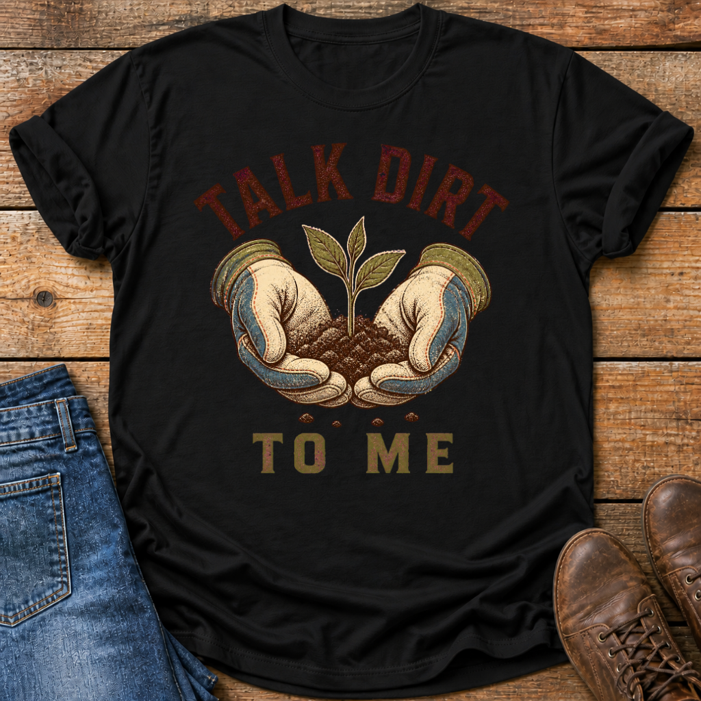 A black graphic t-shirt laid flat on a rustic wooden background, styled with blue jeans and brown boots. The t-shirt features a graphic of two hands holding soil with a small green sprout, with the text 'TALK DIRT TO ME' in dark red and olive green.