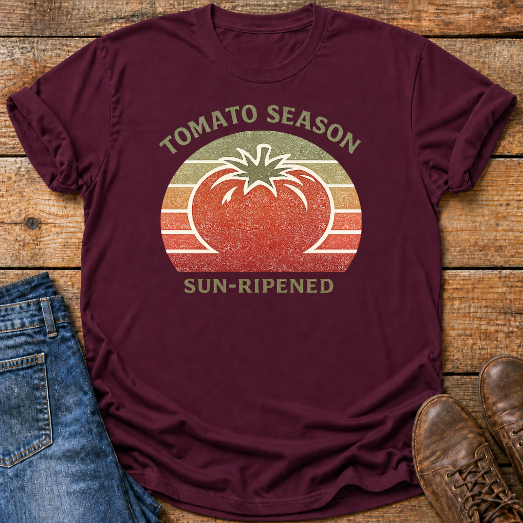 A maroon short-sleeve graphic T-shirt laid flat on a rustic wooden surface, styled with blue jeans on the left and brown shoes on the right. The shirt has a retro circular tomato graphic with the words 'TOMATO SEASON' above and 'SUN-RIPENED' below.