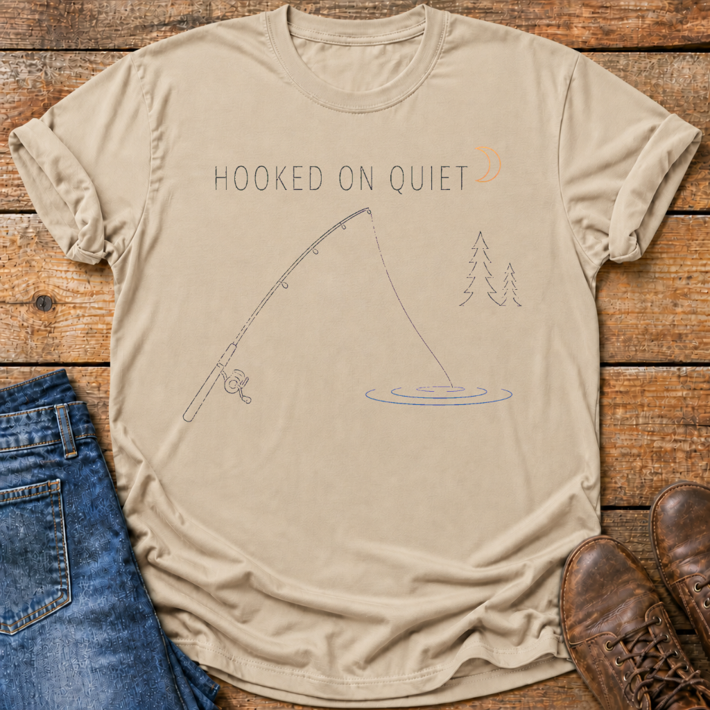A beige graphic t-shirt laid flat on a rustic wooden surface, styled with blue jeans on the left and brown leather shoes on the right. The t-shirt features a graphic of a fishing rod, ripples, pine trees, and the text 'HOOKED ON QUIET' in a muted blue-gray color.