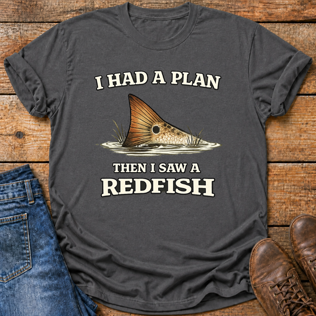 A dark gray graphic t-shirt laid flat on a rustic wooden background, styled with blue jeans on the left and brown leather shoes on the right. The t-shirt features a graphic of a redfish tail and the text 'I HAD A PLAN THEN I SAW A REDFISH' in white and beige lettering.