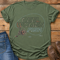 An olive green short-sleeve t-shirt with a 'REEL WOMEN FISH' graphic print, styled in a flat-lay on a rustic wooden background alongside blue jeans and brown leather shoes.