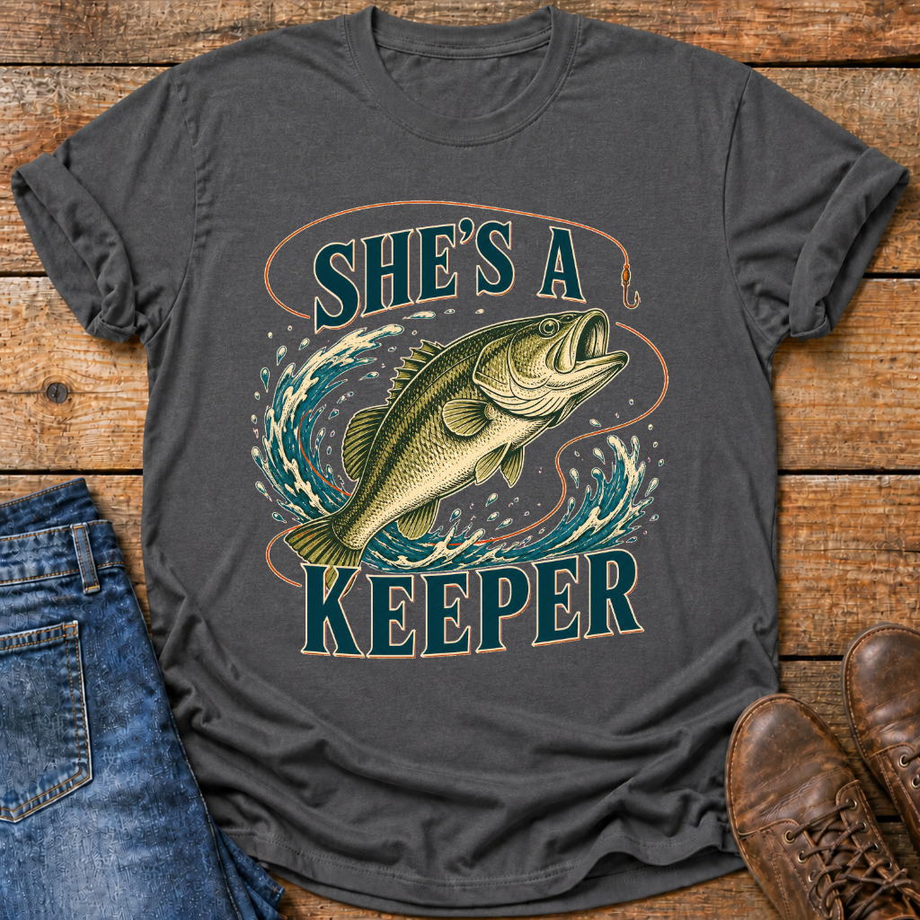 A dark charcoal gray graphic t-shirt featuring a large illustration of a largemouth bass jumping through water with the text 'SHE'S A KEEPER' in teal and tan. The shirt is styled in a flat-lay on a rustic wooden background, partially paired with blue jeans on the left and brown leather shoes on the right.