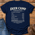 A navy blue graphic t-shirt laid flat on a rustic wooden surface, styled with blue jeans on the left and brown leather shoes on the right. The t-shirt features a humorous 'Deer Camp Nutrition Facts' label printed in white text.