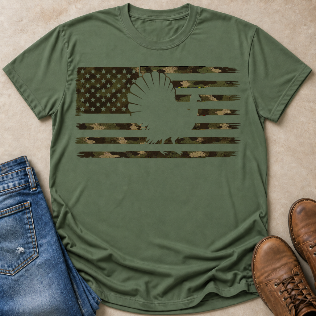 An olive green short-sleeve t-shirt laid flat on a beige textured surface, featuring a distressed camouflage American flag graphic with a turkey silhouette. Blue jeans and brown boots are styled around the shirt.