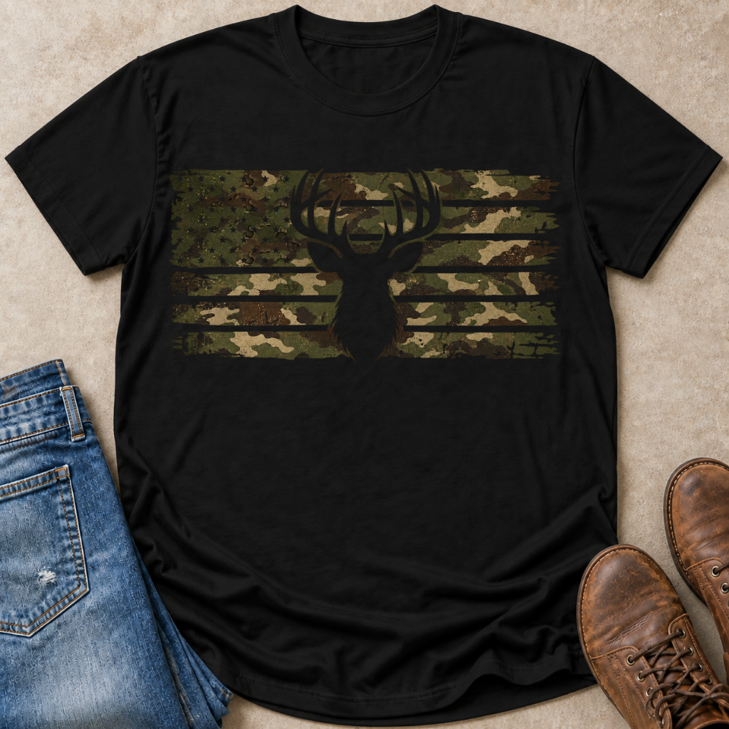 A black short-sleeve t-shirt laid flat on a beige textured surface, featuring a horizontal camouflage american flag graphic across the chest with a deer head silhouette. Blue jeans and brown boots are styled around the shirt.