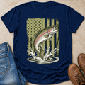 A navy blue graphic t-shirt laid flat on a beige surface, styled with blue jeans on the left and brown leather boots on the right. The t-shirt features a graphic of a jumping rainbow trout in front of a distressed olive green American flag and water splash.