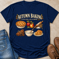 A navy blue graphic t-shirt laid flat on a beige textured surface, styled with blue jeans on the left and brown leather boots on the right. The t-shirt features a 'Autumn Baking' graphic with illustrations of a lattice pie, red apples, a rolling pin, cinnamon sticks, and a bowl of batter.
