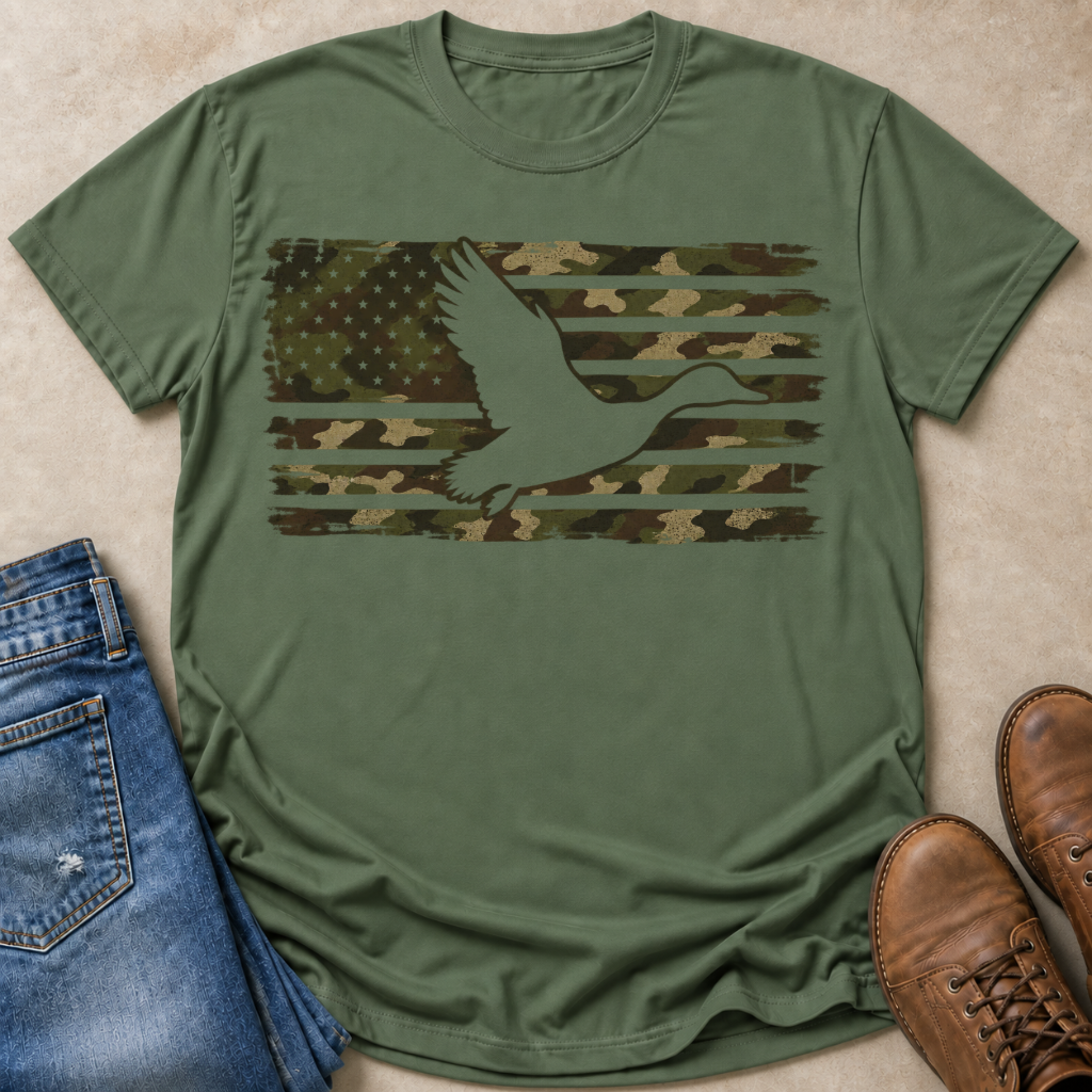 An olive green short-sleeve t-shirt laid flat on a beige surface, featuring a distressed camouflage American flag graphic with a white mallard duck silhouette. Blue jeans and brown leather boots are styled around the shirt.