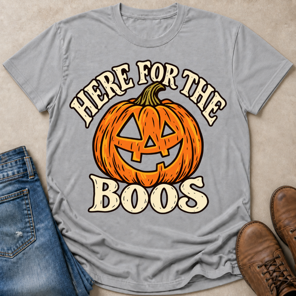 A light gray graphic t-shirt featuring a jack-o'-lantern pumpkin and the text 'HERE FOR THE BOOS' in a distressed cream font. The shirt is styled in a flat-lay arrangement with blue jeans on the left and brown leather boots on the right, set against a neutral beige background.