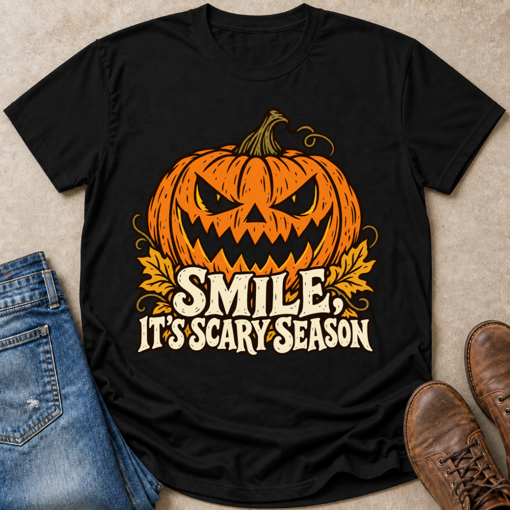 A black short-sleeve graphic T-shirt laid flat on a beige surface, styled with blue jeans, brown shoes, and a small amount of blue pen in the corners. The shirt features a large orange jack-o'-lantern face with a carved expression and the phrase “SMILE, IT’S SCARY SEASON” in cream and orange lettering.