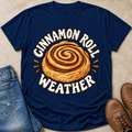 A navy blue graphic t-shirt laid flat on a beige surface, styled with blue jeans on the left and brown boots on the right. The t-shirt features a large graphic of a cinnamon roll with the text 'CINNAMON ROLL WEATHER' in white arched above and below.