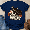 A navy blue graphic t-shirt laid flat on a beige textured surface, styled with blue jeans on the left and brown boots on the right. The t-shirt features a graphic of a black cat with yellow eyes resting on a mug, surrounded by orange autumn leaves and the text 'COZY WITH A CHANCE OF MISCHIEF' in cream and white.